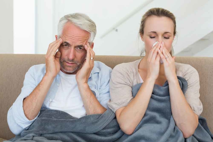 Two people sitting on a couch, one with a headache, the other with a stuffy nose